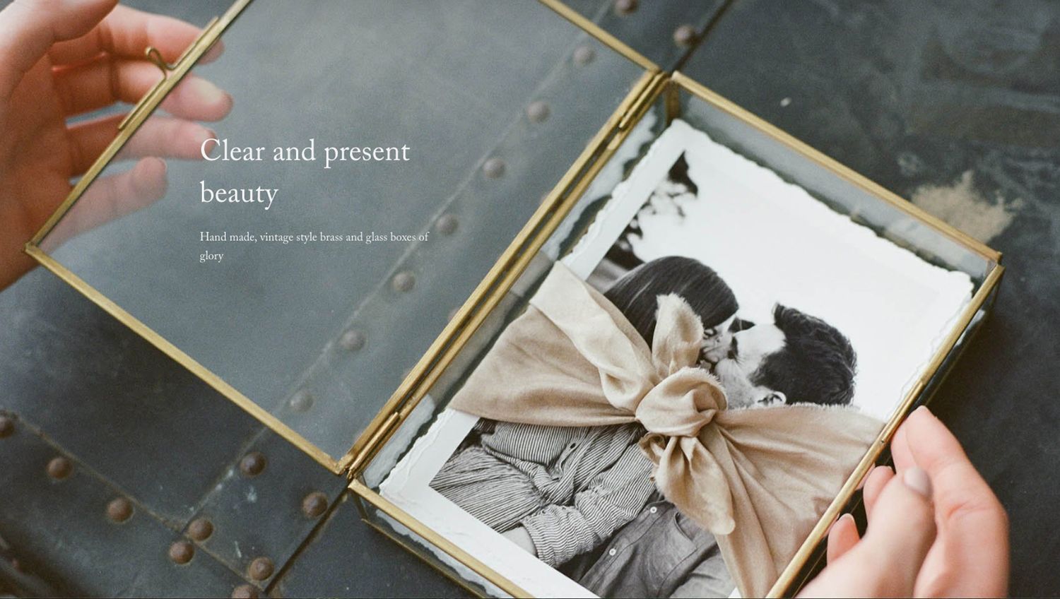 Hands holding an open gold-framed photo book displaying a vintage black-and-white romantic photograph tied with a silk bow.