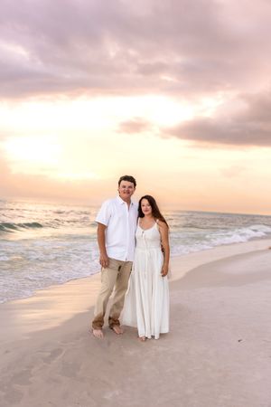 Couple posing together on sandy beach at sunset, man in white shirt and khakis, woman in white dress.