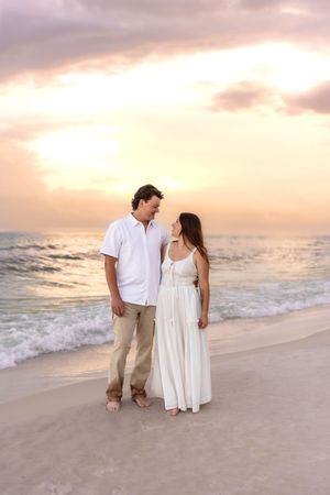 Couple standing together on beach at sunset, woman in white maxi dress, man in white shirt.