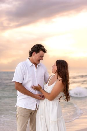 Couple smiling at each other on a beach at sunset, man in white shirt and woman in white dress.