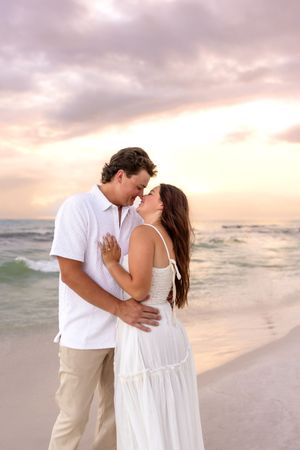 Romantic couple nearly kissing on a beach at sunset, embracing in white coordinated outfits.
