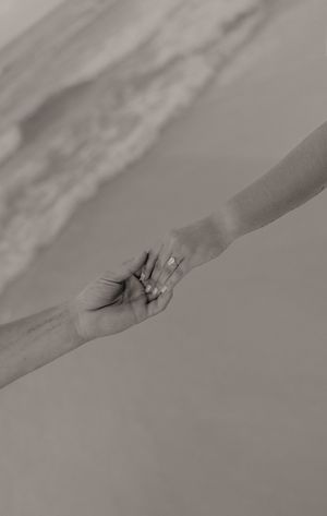 Black and white close-up of couple holding hands with engagement ring on beach.