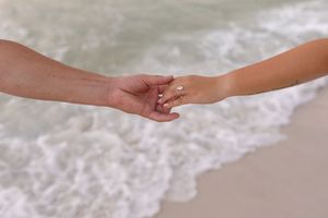 Close-up of couple holding hands on a sandy beach with gentle ocean waves in background.