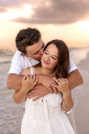 Man kissing woman on cheek from behind at beach sunset, couple embracing in white outfits.