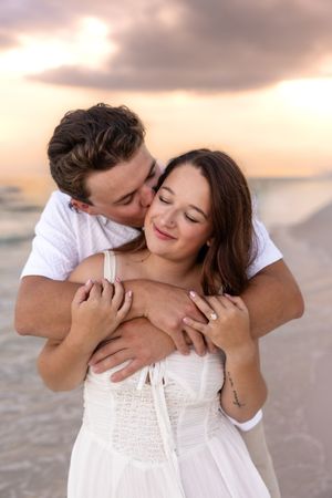 Man kissing woman on cheek from behind while embracing her on a beach at sunset.