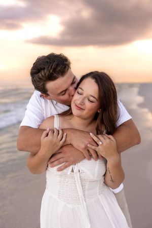Couple embracing at beach sunset, man kissing woman's cheek while hugging her from behind, both wearing white.