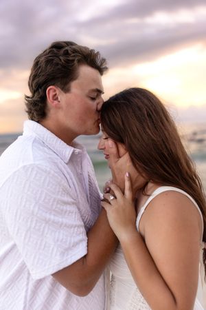 Romantic couple sharing a kiss on the beach at sunset, woman wearing engagement ring, pastel sky background.