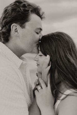 Black and white close-up of couple sharing a tender kiss on the cheek at the beach.
