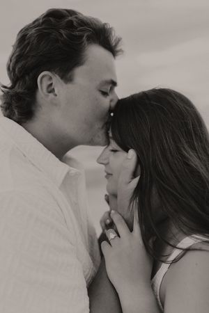 Black and white close-up of couple sharing a tender kiss on forehead at the beach.