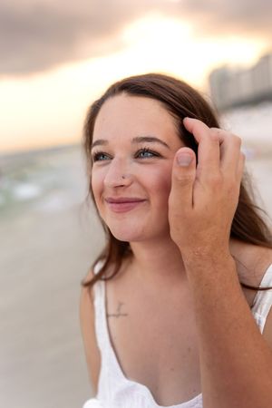 Smiling young woman with blue eyes on a beach at sunset, hand gently touching her face.