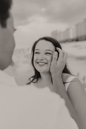 Black and white photo of smiling woman as man gently touches her face on a beach, buildings visible in background.