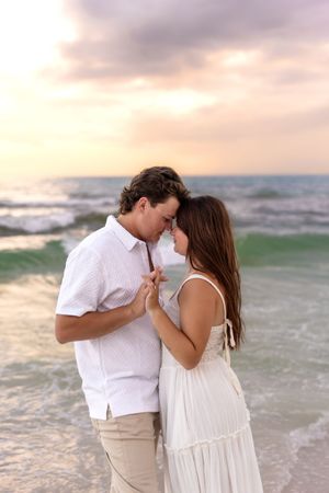 Romantic couple in white outfits touching foreheads tenderly on a beach at sunset with ocean waves behind them.