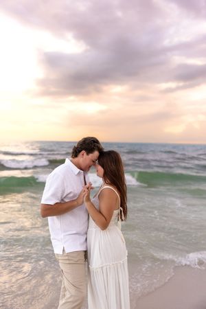Couple touching foreheads romantically on a beach at sunset, holding hands in white outfits.