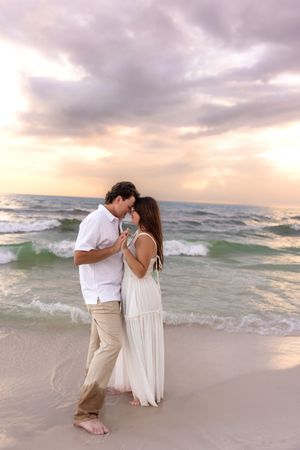 Couple sharing intimate moment on beach at sunset, holding hands with ocean waves behind them.