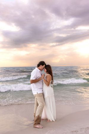 Romantic couple embracing on a beach at sunset with ocean waves and dramatic cloudy sky behind them.