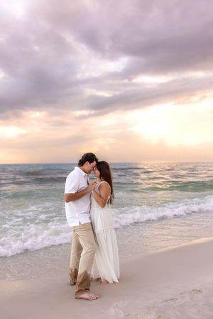 Romantic couple slow dancing and embracing on a sandy beach at sunset with ocean waves.