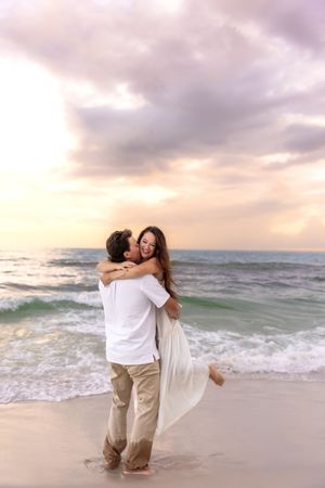 Happy couple embracing and laughing on a sandy beach at sunset, man lifting woman in a flowing white dress.