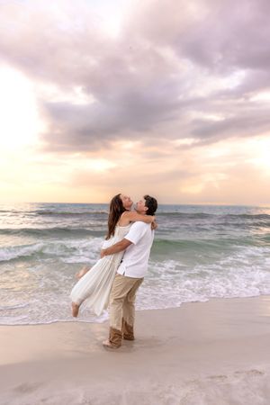 Couple sharing a romantic kiss on a beach at sunset, woman lifted in mans arms with ocean waves behind them.
