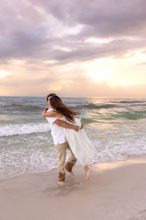 Couple embracing on a beach at sunset, waves crashing behind them, woman in white dress lifted by man in white shirt.