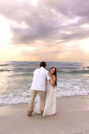 Couple embracing and laughing together on sandy beach during vibrant pink and gold sunset.