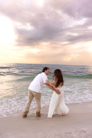 Couple dancing and laughing together on a beach at sunset with waves crashing behind them.