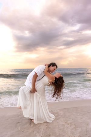 Man dipping woman in white dress dramatically on a beach at sunset with colorful sky.