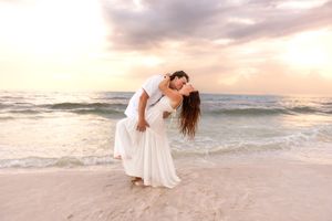 Couple sharing a romantic dip kiss on sandy beach with colorful sunset sky backdrop.