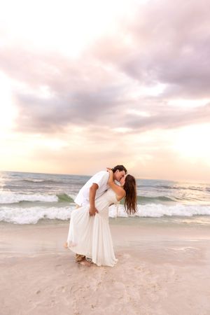 Couple sharing a passionate dip kiss on a sandy beach during a colorful sunset.