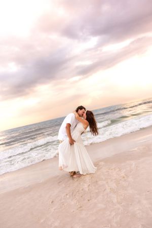 Couple sharing a passionate kiss on a sandy beach at sunset, both dressed in white, ocean waves behind them.