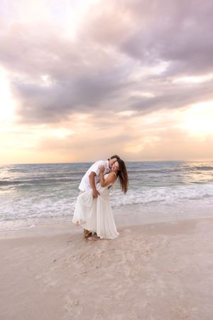 Couple embracing and dipping on a sandy beach with dramatic purple sunset sky behind them.