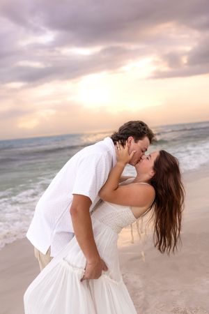 Couple sharing a romantic kiss while embracing on a sandy beach during a colorful sunset.