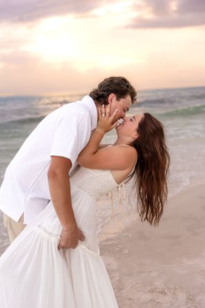 Couple sharing a romantic kiss on beach at sunset, woman in flowing white dress.
