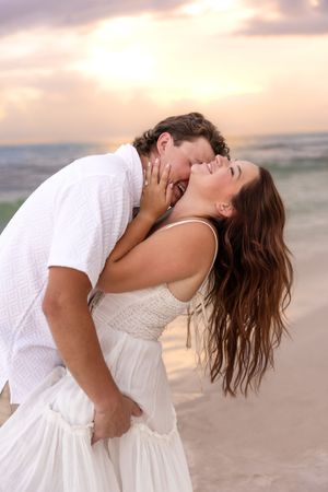 Couple sharing a playful kiss on a sandy beach at sunset, woman laughing with hair flowing.