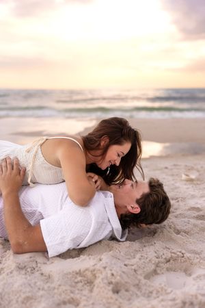 Playful couple lying on beach sand laughing together during a colorful sunset by the ocean.