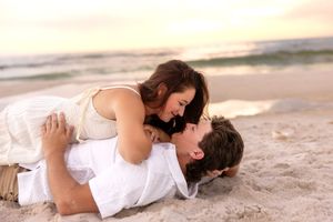 Romantic couple lying on sandy beach nearly kissing, woman in white dress leaning over man at sunset.