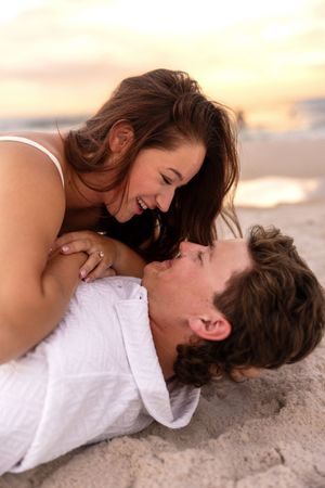 Couple smiling and gazing at each other while lying on sandy beach during a golden sunset.