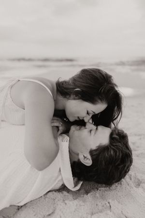 Black and white photo of romantic couple almost kissing while lying on sandy beach in intimate moment.