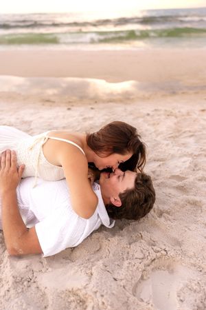 Romantic couple lying on sandy beach sharing a kiss at sunset with gentle ocean waves in background.