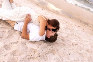 Romantic couple lying together on sandy beach, sharing a kiss near the ocean shoreline.