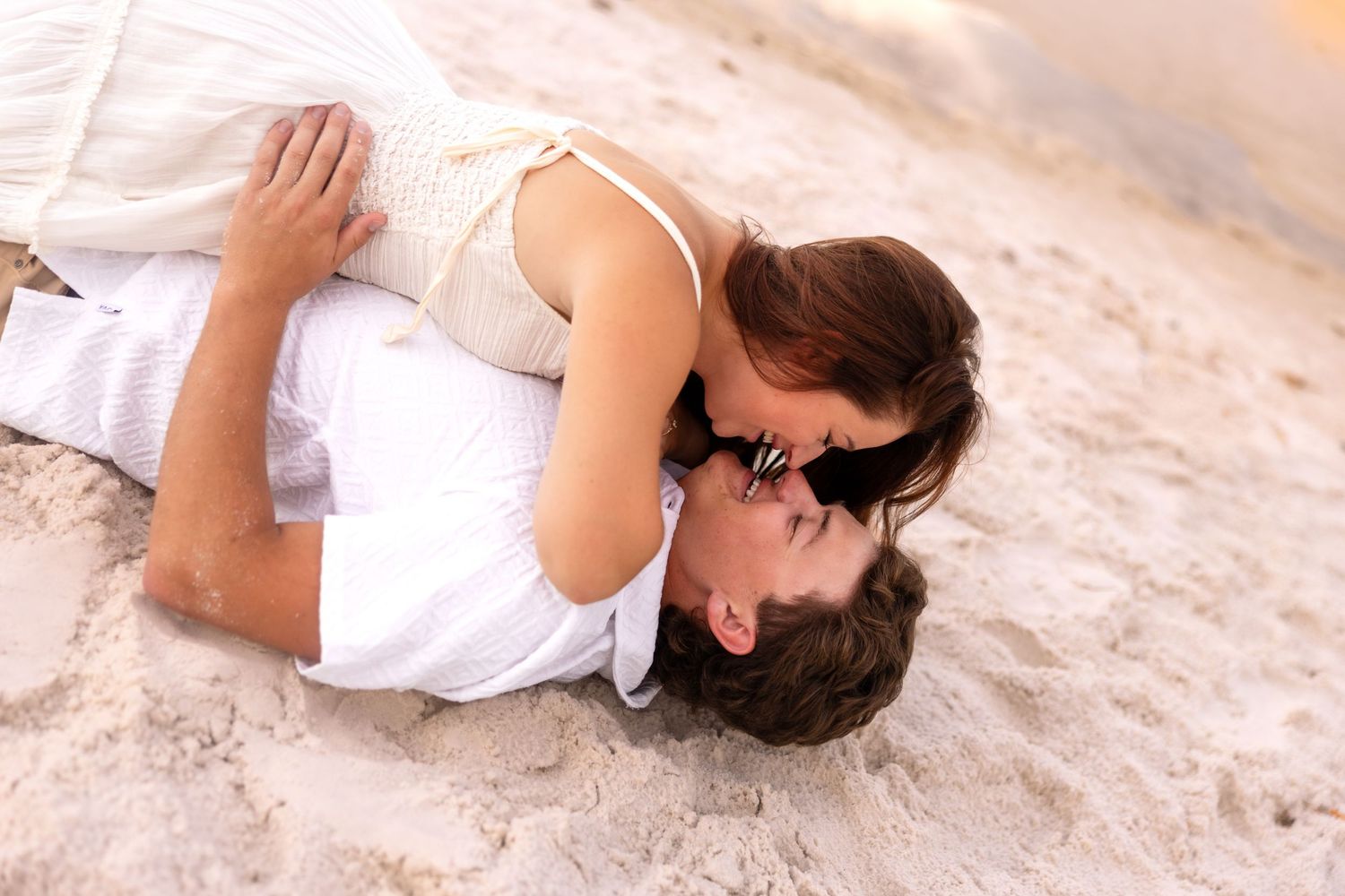 Romantic couple sharing a tender kiss while lying on sandy beach in white outfits.