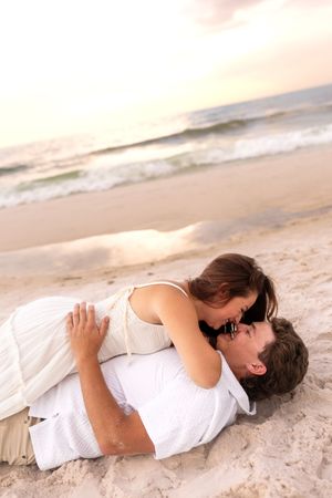 Couple lying on sandy beach sharing a romantic moment, woman in white dress leaning over man.