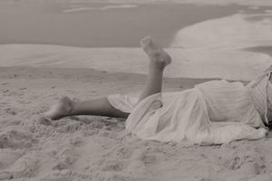 Black and white photo of a woman in a white dress lying on a sandy beach with legs raised.