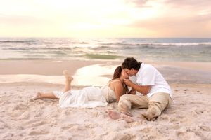 Romantic couple sharing a tender kiss on a sandy beach at sunset, with gentle ocean waves in the background.