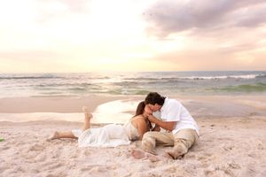 Couple kissing while lying on sandy beach at sunset with pastel sky and ocean waves.