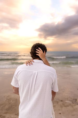 Woman's hand resting on man's shoulder and head as they face a colorful beach sunset.
