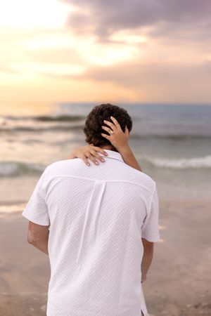 Close-up of woman's hand on man's shoulder and hair while facing a pastel beach sunset.