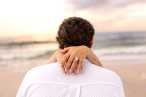 Woman with engagement ring embracing man from behind on beach at sunset.