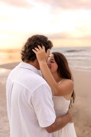 Couple sharing a tender kiss on the beach at sunset, woman in white dress embracing man.