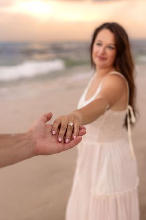 Close-up of woman showing engagement ring while holding partner's hand on beach.