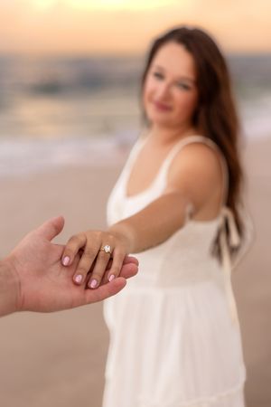 Woman in white dress showing engagement ring on beach at sunset.